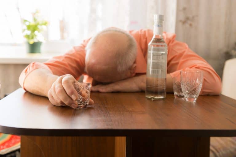 close up velho careca bebado encostado na mesa de madeira com vodka segurando um copo pequeno